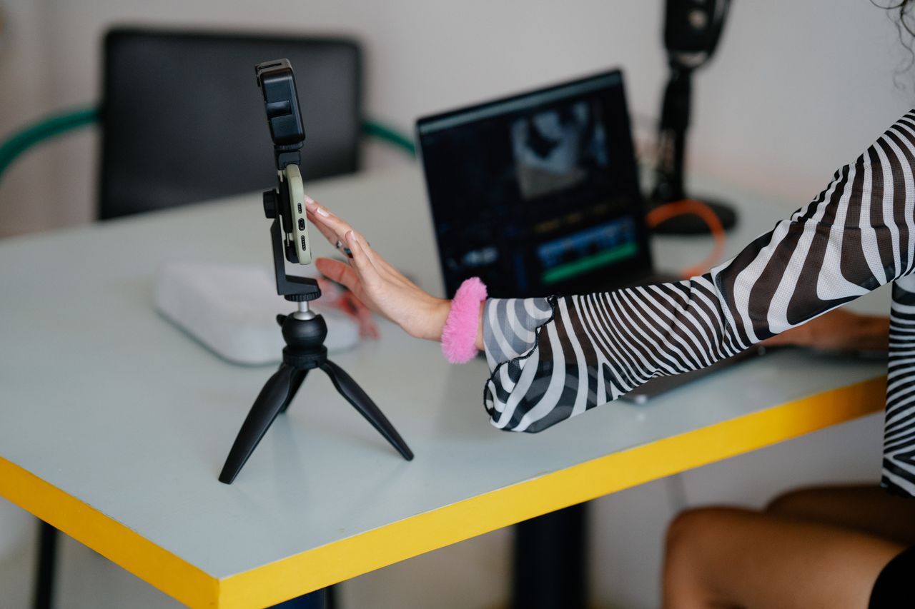 services-02 Woman adjusting smartphone on tripod with laptop setup for video editing.